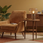 A cognac ribbed Clérachic lamp on a round wooden side table beside a tan velvet armchair, with a large green plant behind.