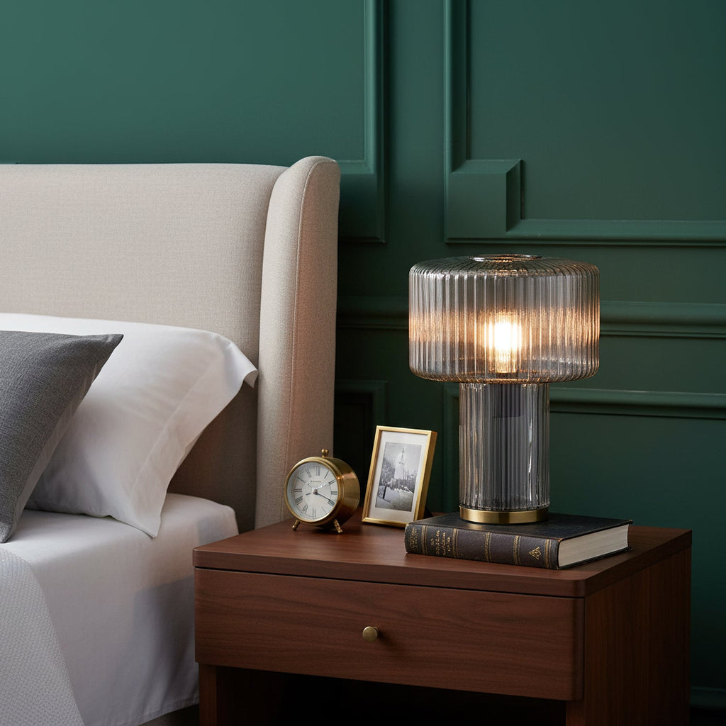 Smoky grey ribbed glass Clérachic lamp on a walnut bedside table with a brass clock, a framed photo and a book, against green panelling.