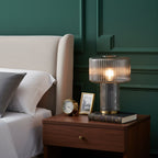 Smoky grey ribbed glass Clérachic lamp on a walnut bedside table with a brass clock, a framed photo and a book, against green panelling.