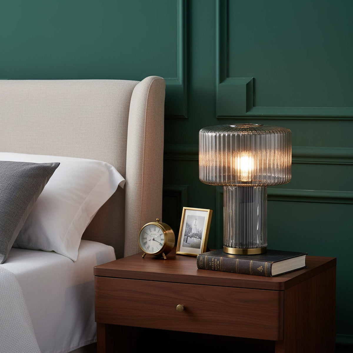 Smoky grey ribbed glass Clérachic lamp on a walnut bedside table with a brass clock, a framed photo and a book, against green panelling.
