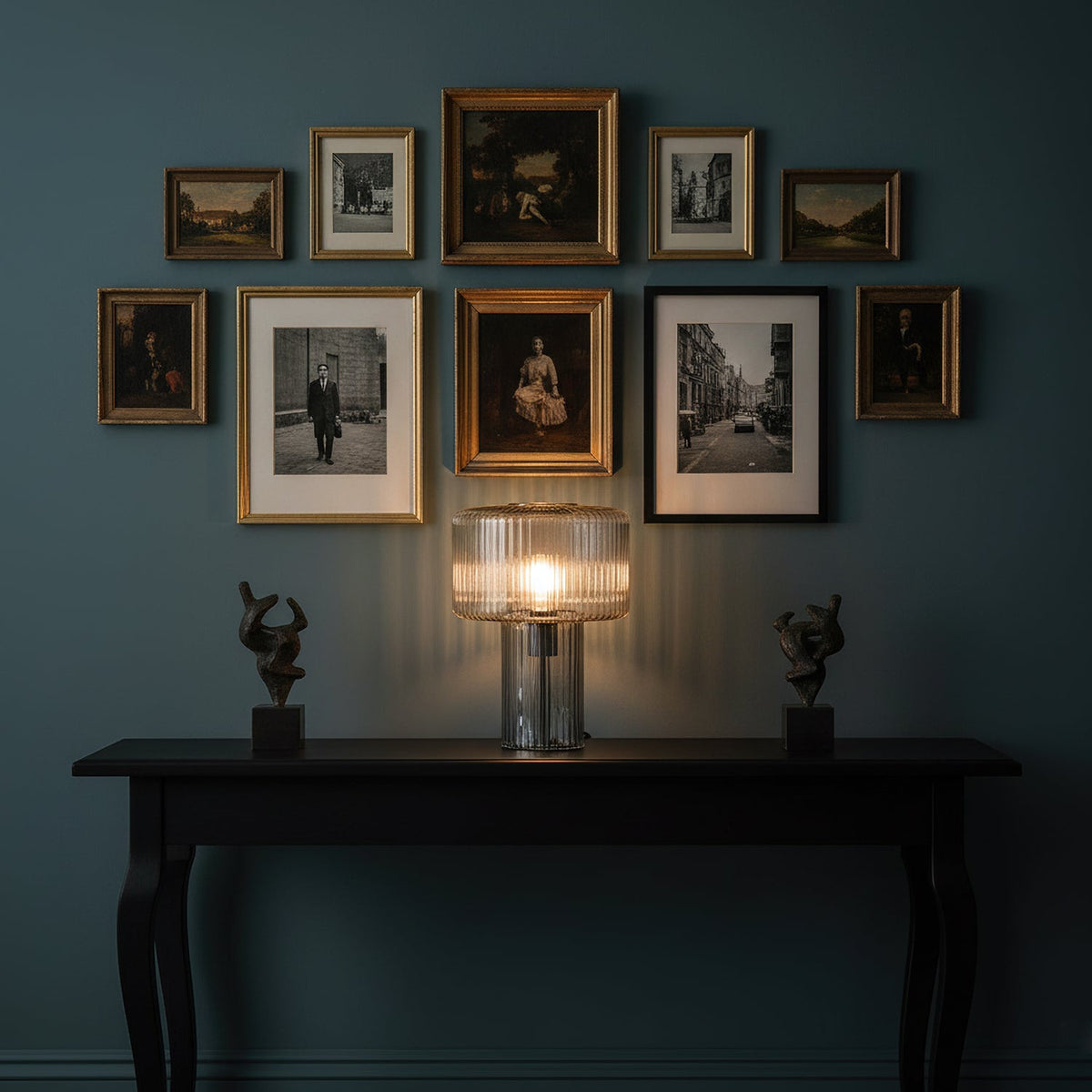Lit ribbed glass Clérachic lamp on a black console table, under a gathered wall of framed prints and flanked by two small figures.