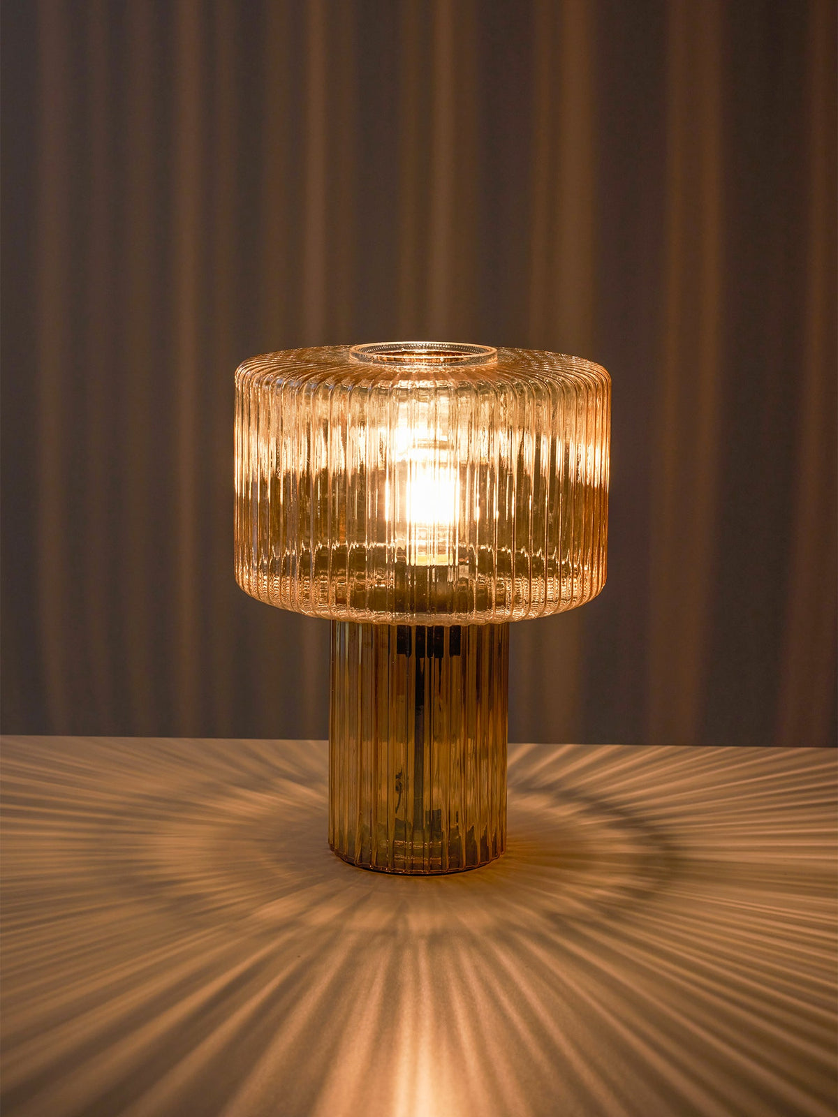 Cognac ribbed glass Clérachic lamp glowing on a tabletop, casting a starburst of light onto the surface and a curtain behind.