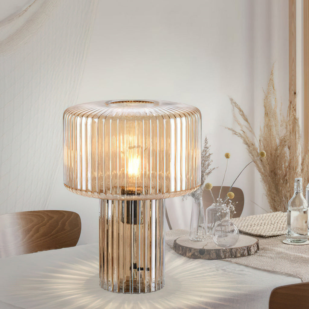 Large cognac ribbed glass Clérachic lamp on a white dining table, with a wood slice, glass bottles and dried pampas grass behind.