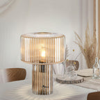 Large cognac ribbed glass Clérachic lamp on a white dining table, with a wood slice, glass bottles and dried pampas grass behind.