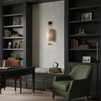 A Clérachic ribbed wall light glowing softly in an alcove between dark built-in bookshelves, beside a green armchair and a black desk.