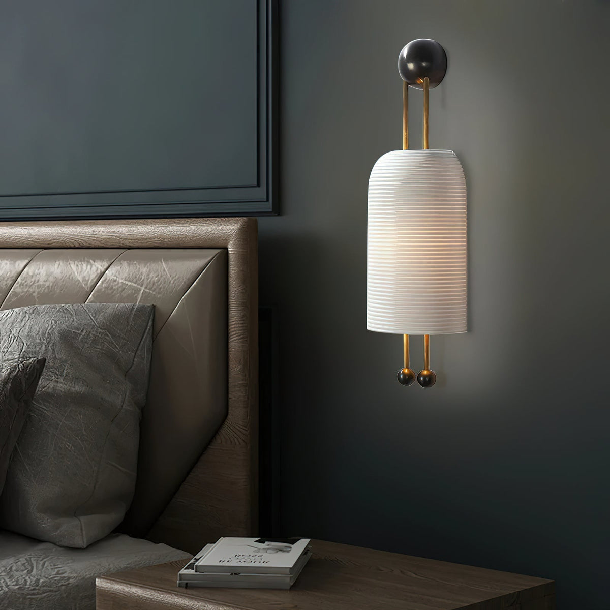 Close side view of a ribbed white Clérachic wall light on a dark grey-green wall above a leather headboard and a stack of books.