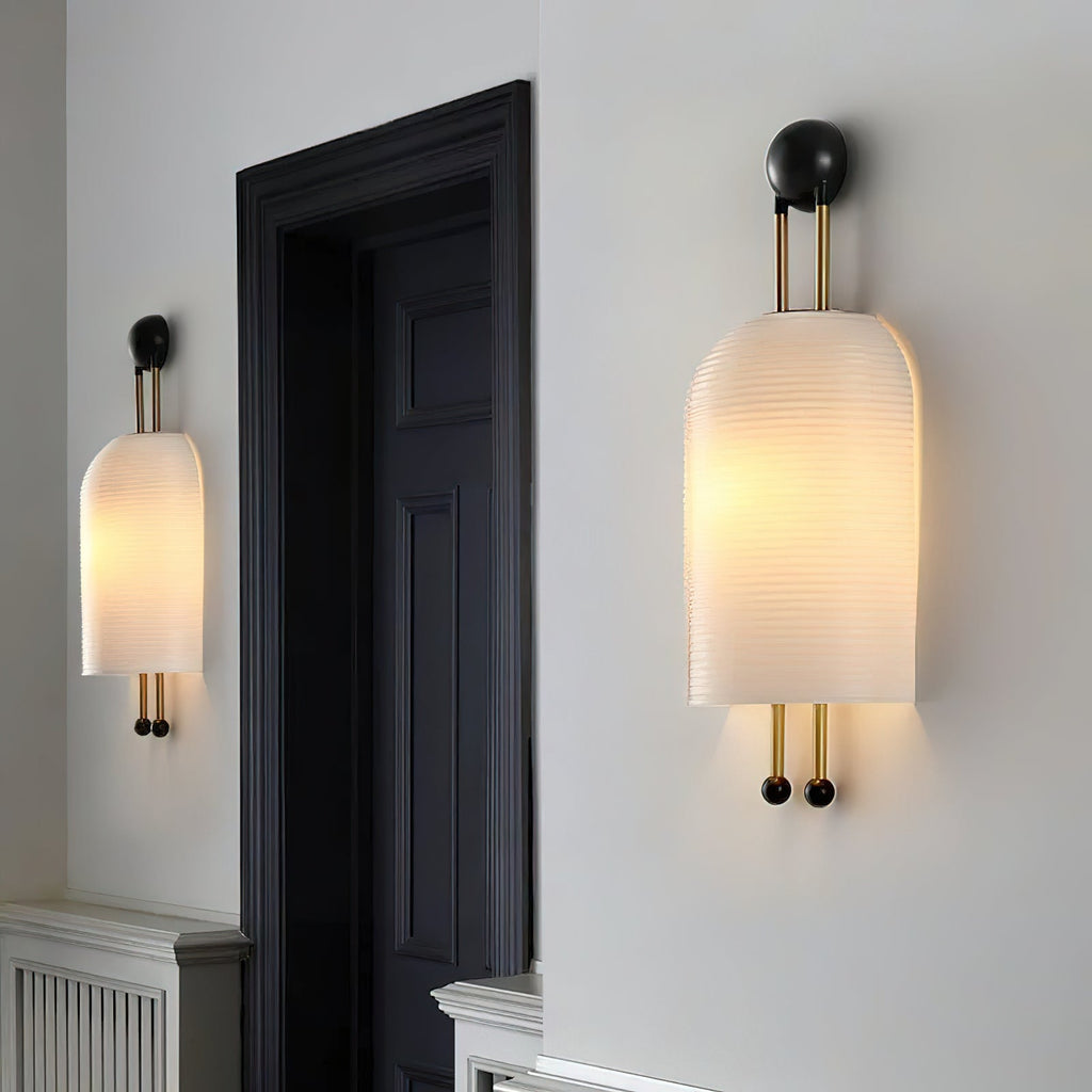 Two Clérachic ribbed wall lights lit on a pale wall beside a black door frame, one in the foreground and one further along the wall.