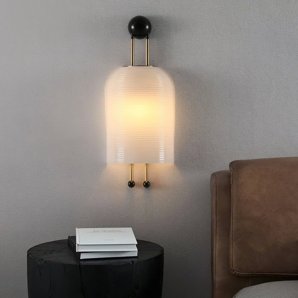Lit ribbed white shade of this Clérachic wall light on a grey wall above a round black side table and a brown leather headboard.