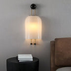 Lit ribbed white shade of this Clérachic wall light on a grey wall above a round black side table and a brown leather headboard.