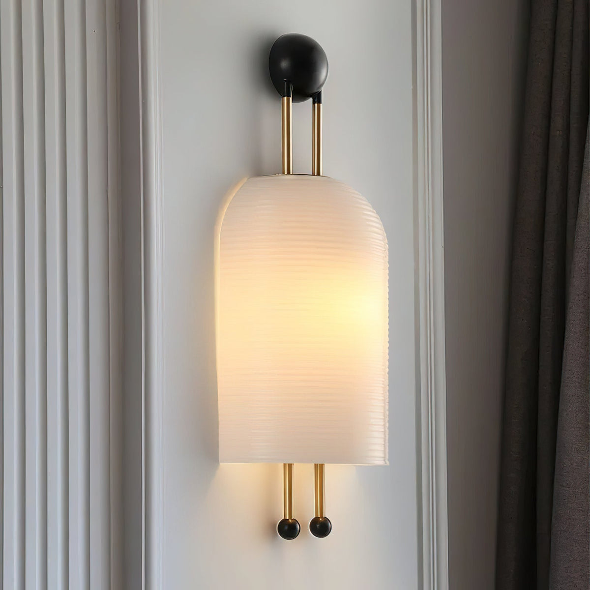 Large ribbed white Clérachic wall light glowing on a white panelled wall, with its gold rods and black ball ends beside a dark curtain.