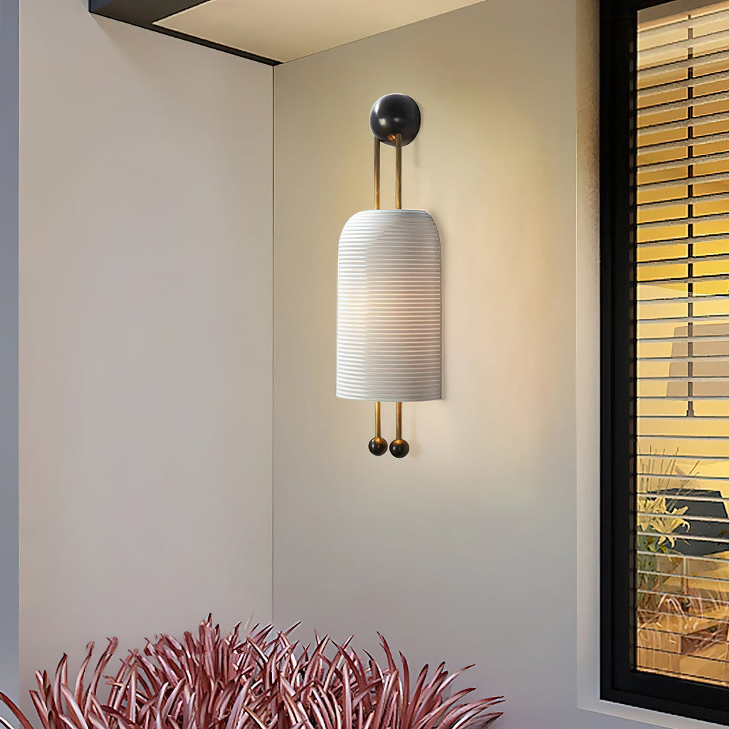A Clérachic wall light with a ribbed white shade on a cream wall beside a black-framed window, with spiky red-pink leaves at the bottom.