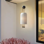 A Clérachic wall light with a ribbed white shade on a cream wall beside a black-framed window, with spiky red-pink leaves at the bottom.