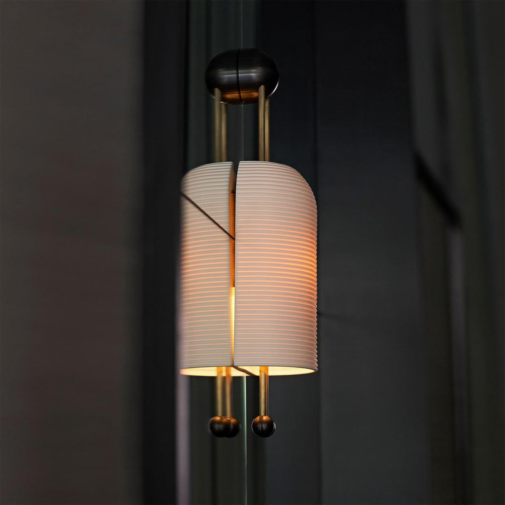 Lit ribbed shade of this Clérachic wall light against a dark curtain, showing the gold rods, the black wall plate and the two ball ends.