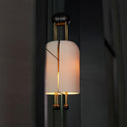 Lit ribbed shade of this Clérachic wall light against a dark curtain, showing the gold rods, the black wall plate and the two ball ends.