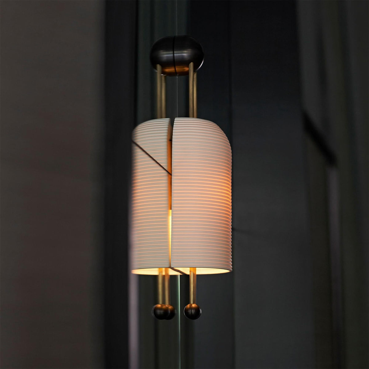 Lit ribbed shade of this Clérachic wall light against a dark curtain, showing the gold rods, the black wall plate and the two ball ends.