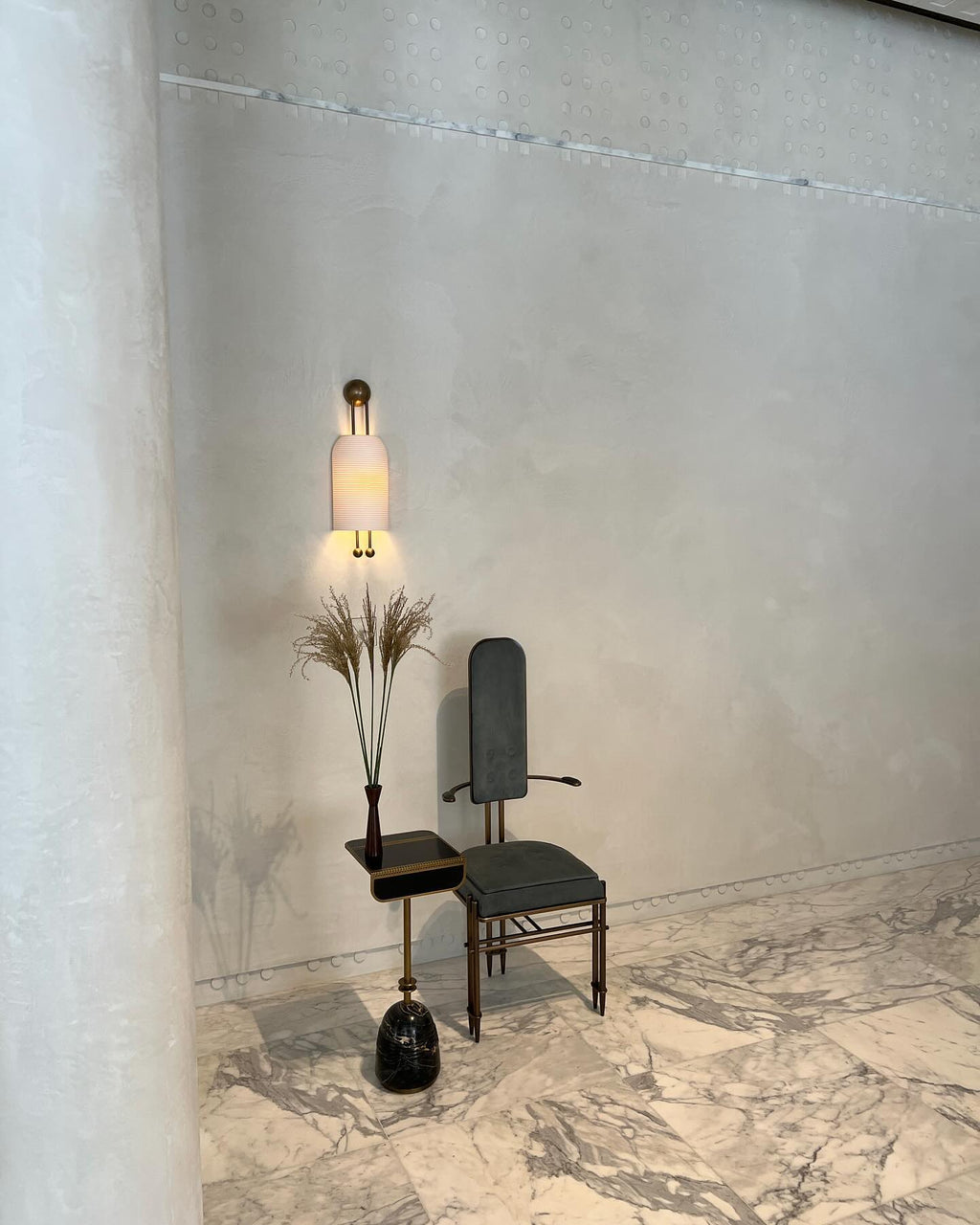 Lit Clérachic wall sconce on a plaster-finish wall above a small dark chair and side table with a tall dried-stem arrangement.