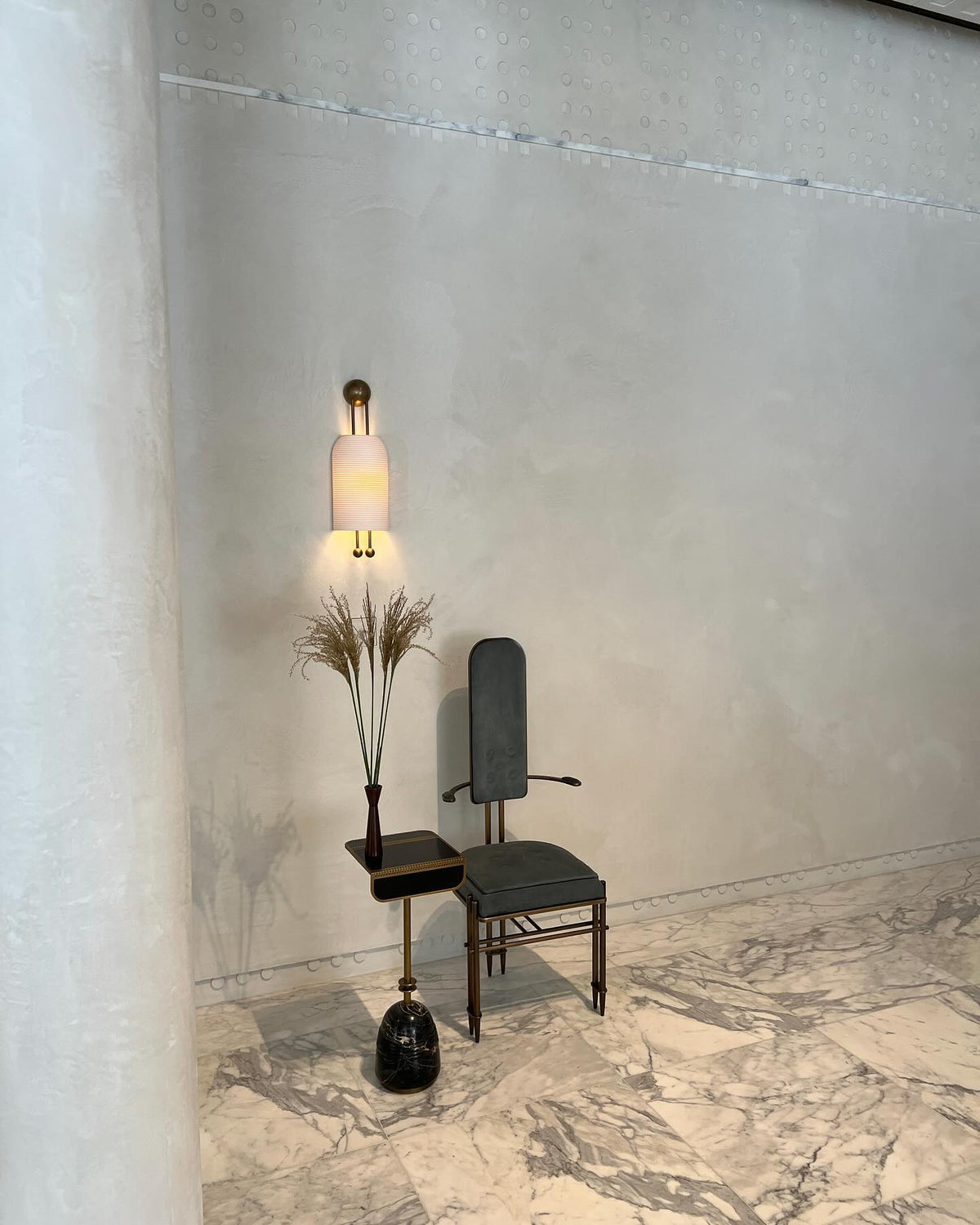 Lit Clérachic wall sconce on a plaster-finish wall above a small dark chair and side table with a tall dried-stem arrangement.