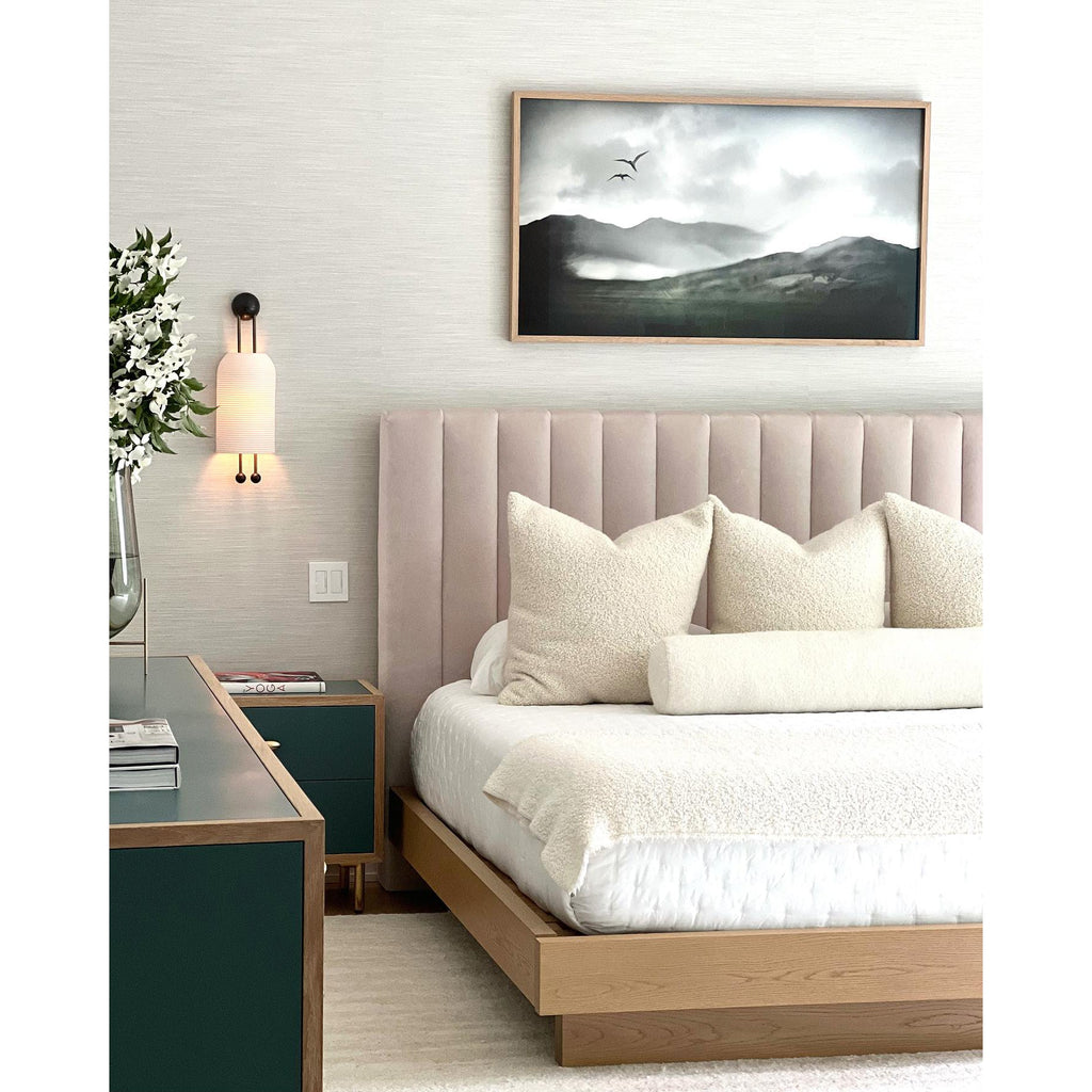 Clérachic wall light with a ribbed white shade beside a blush pink channel-tufted headboard, with white bedding and a framed landscape print.