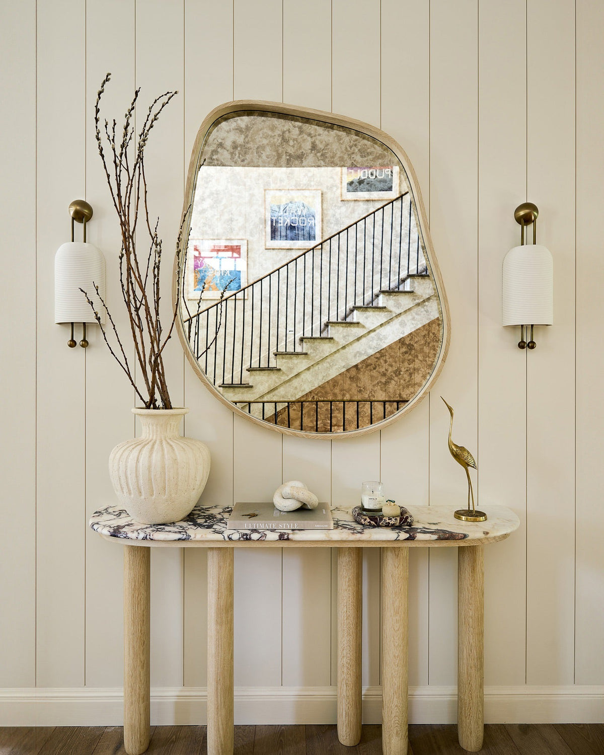 Pair of Clérachic ribbed wall lights either side of a curved-edge mirror above a marble-top console with a vase of bare branches.