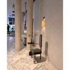 A lit Clérachic wall light on a pale plaster wall in a marble-floored showroom, with a dark chair and slim round-based side table below it.