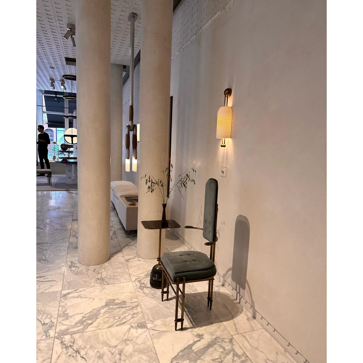 A lit Clérachic wall light on a pale plaster wall in a marble-floored showroom, with a dark chair and slim round-based side table below it.