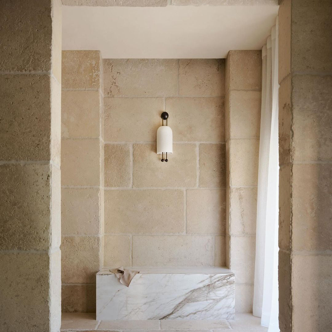 A small Clérachic wall light on a sandstone block wall above a marble bench, with a sheer curtain at the right edge of the alcove.
