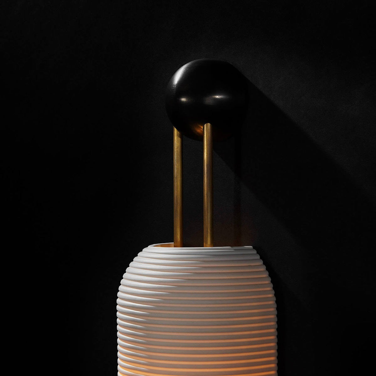Close view of the black round wall plate, gold rods and ribbed top of the shade on this Clérachic wall light, against a black backdrop.