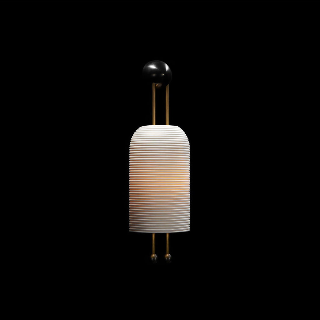 This Clérachic ribbed wall light, softly lit and set against a plain black backdrop, with its black wall plate and gold rods above the shade.