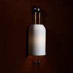 Lit ribbed white shade of this Clérachic wall light on a deep brown wall, with its gold rods and black ball ends in warm shadow.