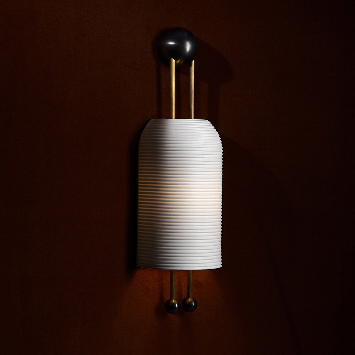 Lit ribbed white shade of this Clérachic wall light on a deep brown wall, with its gold rods and black ball ends in warm shadow.