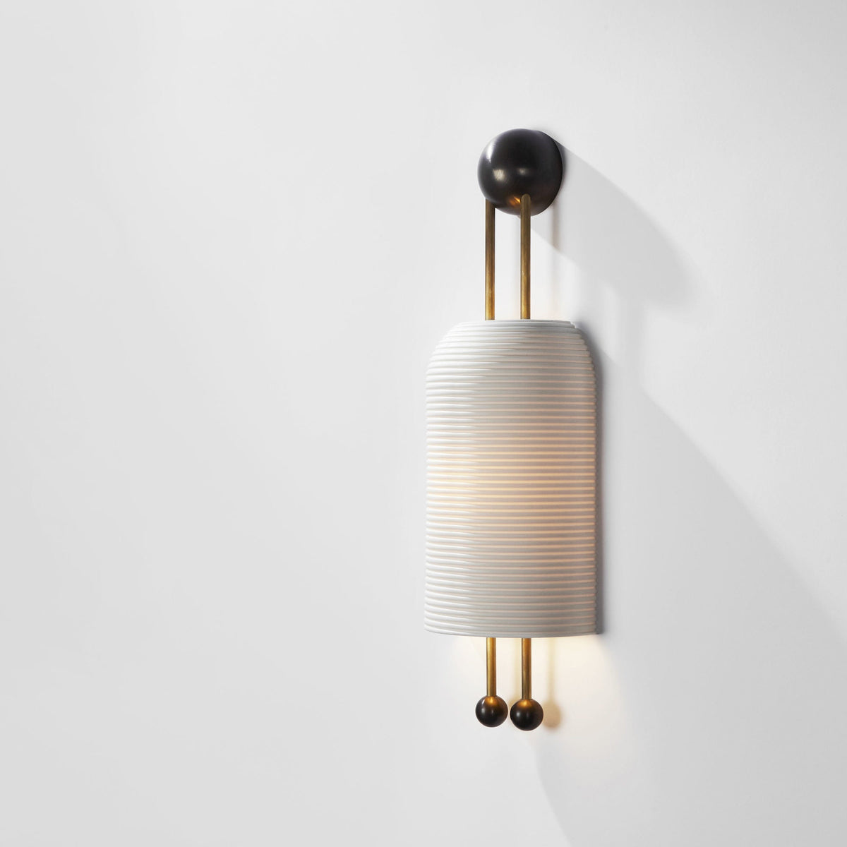 A lit Clérachic ribbed wall light casting a gentle shadow on a bright white wall, with a black wall plate and black ball ends.