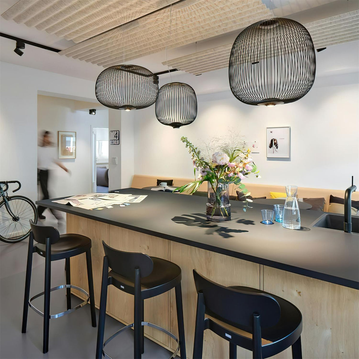 Three black wire-cage pendants from Clérachic hang at different heights over a long black-topped island with three black bar stools.