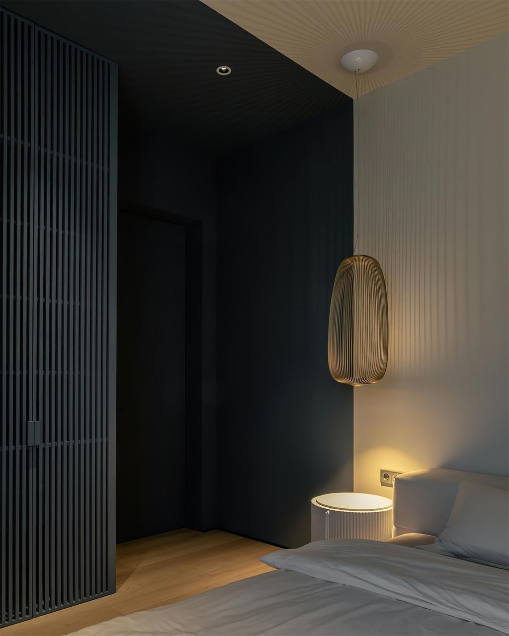 A single tall black wire-cage Clérachic pendant glows beside a bed in a dark bedroom with a slatted black wall panel.