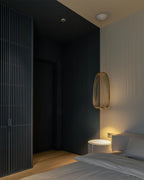 A single tall black wire-cage Clérachic pendant glows beside a bed in a dark bedroom with a slatted black wall panel.