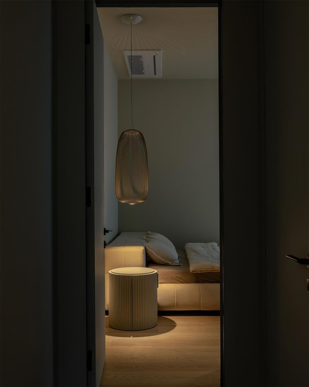 Seen through a dark doorway, a tall Clérachic wire-cage pendant lights a small sitting corner with a low stool and a day bed.
