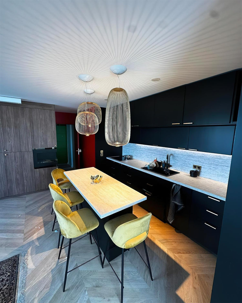 A cluster of Clérachic wire-cage pendants hangs over a narrow island with yellow stools, with a black kitchen along the wall.
