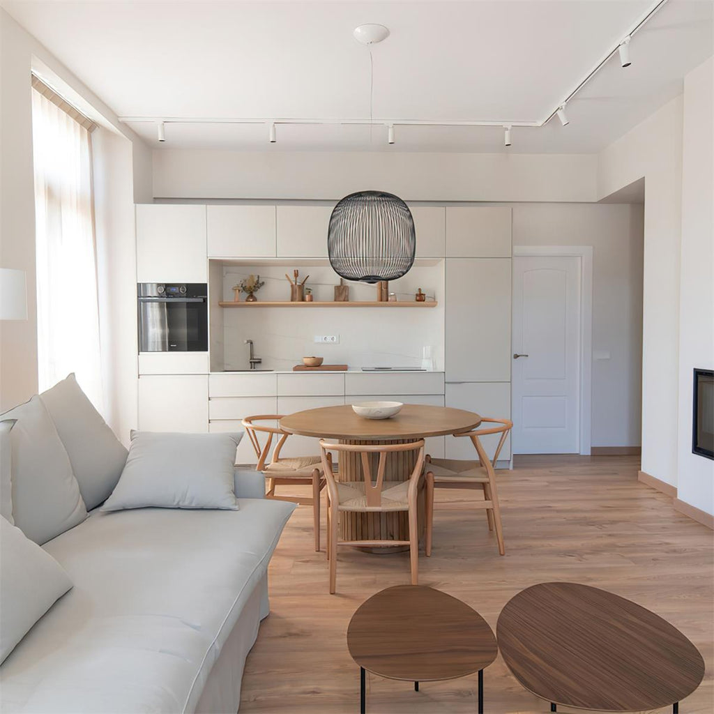 A round wire-cage Clérachic pendant hangs in an open-plan living and kitchen space, above a round table and wooden chairs.