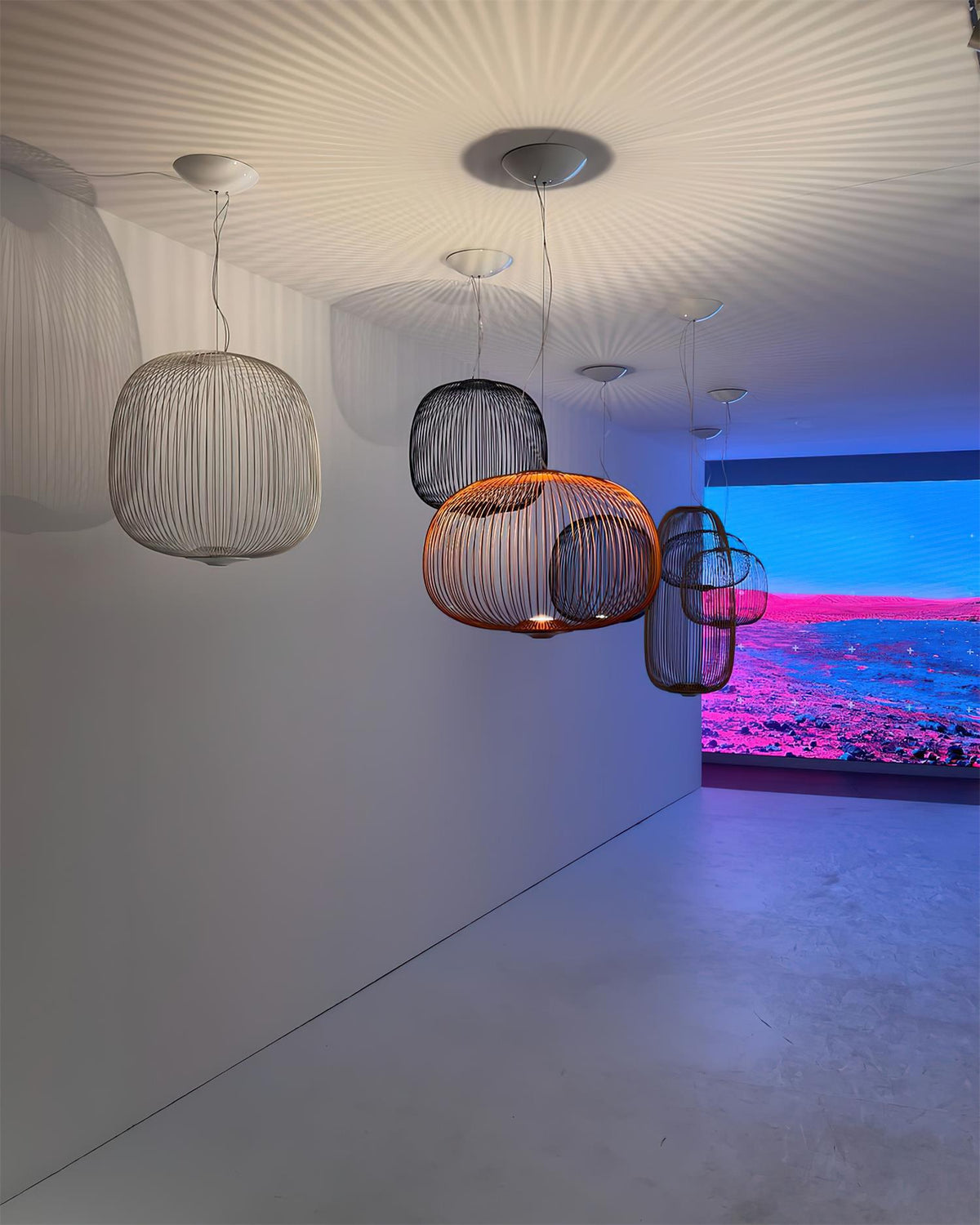 Three Clérachic wire-cage pendants in grey and a copper tone hang in a grey room with a bright pink and blue screen behind.