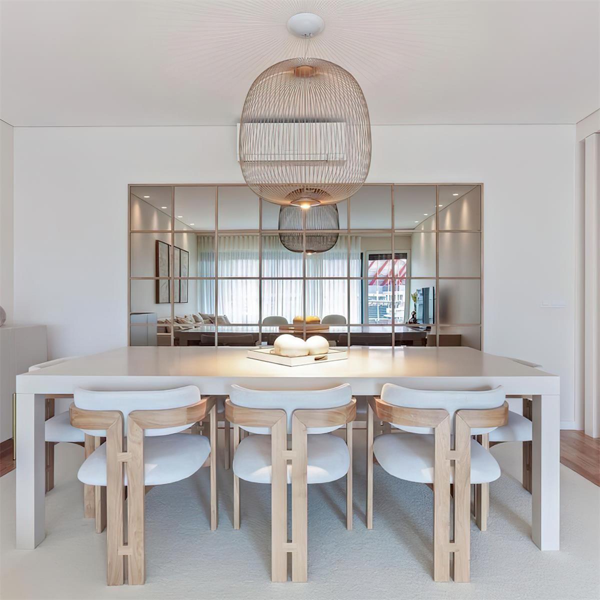 A large gold-toned Clérachic wire-cage pendant hangs over a white dining table with six white chairs, with a mirrored wall behind.