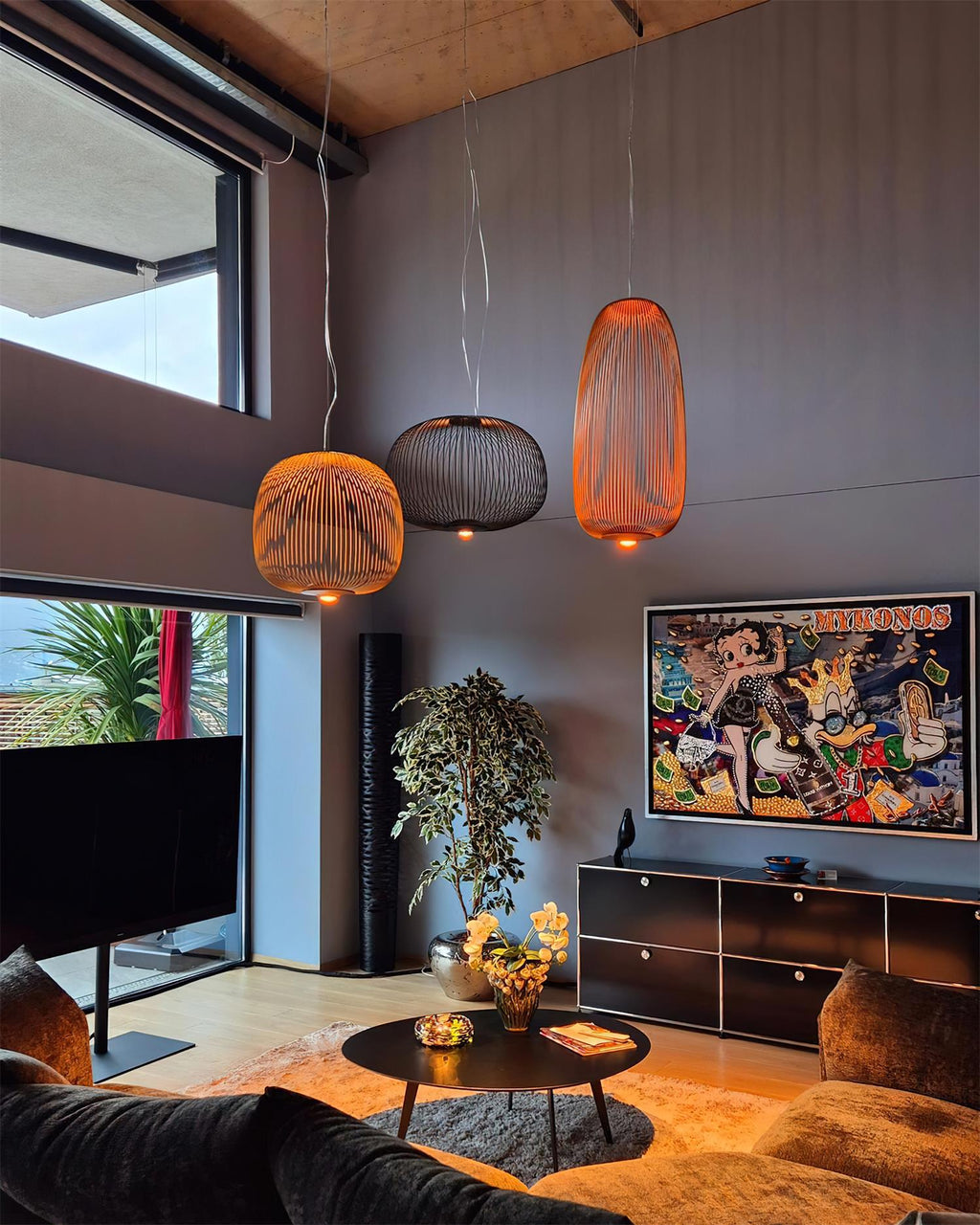 Three Clérachic wire-cage pendants in copper, black and orange tones hang over a living room with a dark sofa and a colourful artwork.