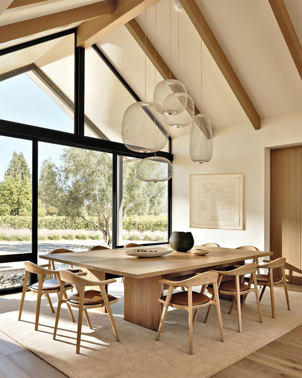 Several white wire-cage Clérachic pendants hang under a beamed ceiling with large windows, over a long wooden dining table.