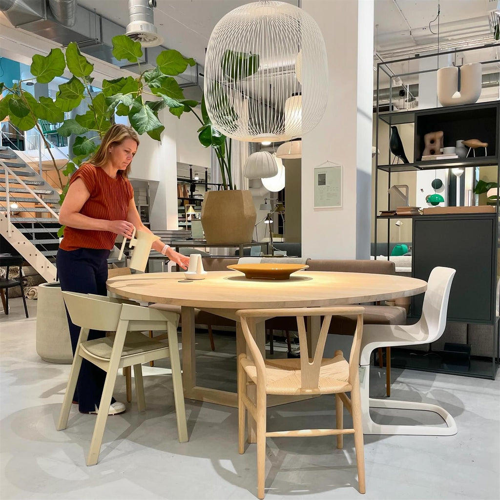 A woman sets a round table below large white Clérachic wire-cage pendants in a bright showroom with a big leafy plant.