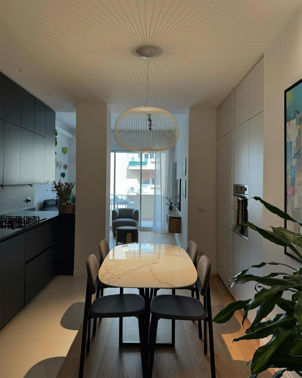 A wide white wire-cage Clérachic pendant hangs in a narrow room above a long oval dining table, with a dark kitchen at its side.