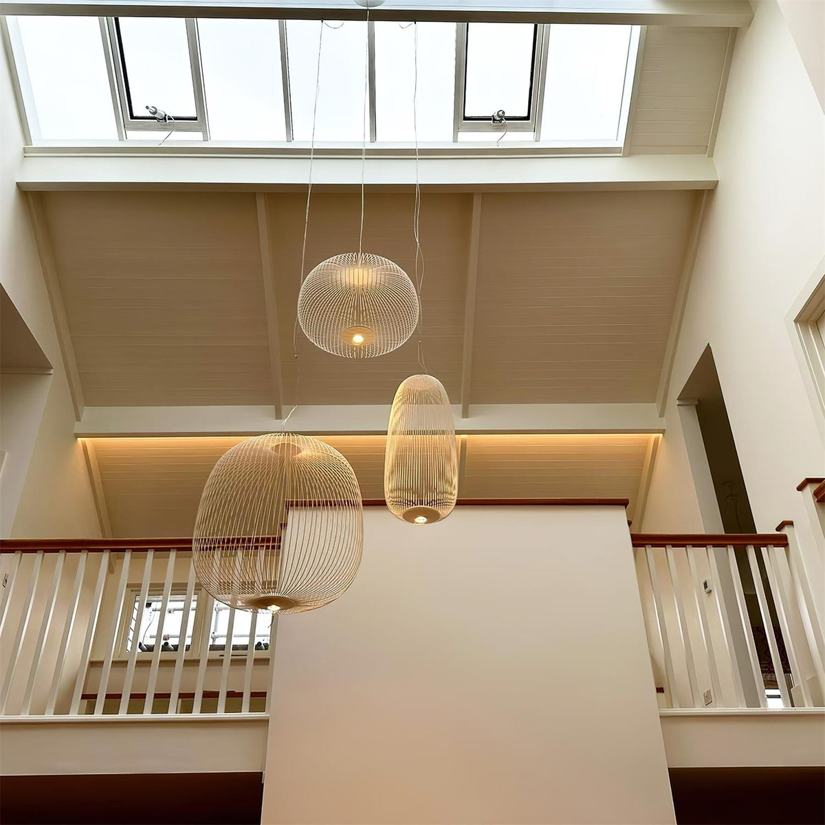 Two white wire-cage Clérachic pendants hang at different heights in a high space with a skylight and a white balustrade.