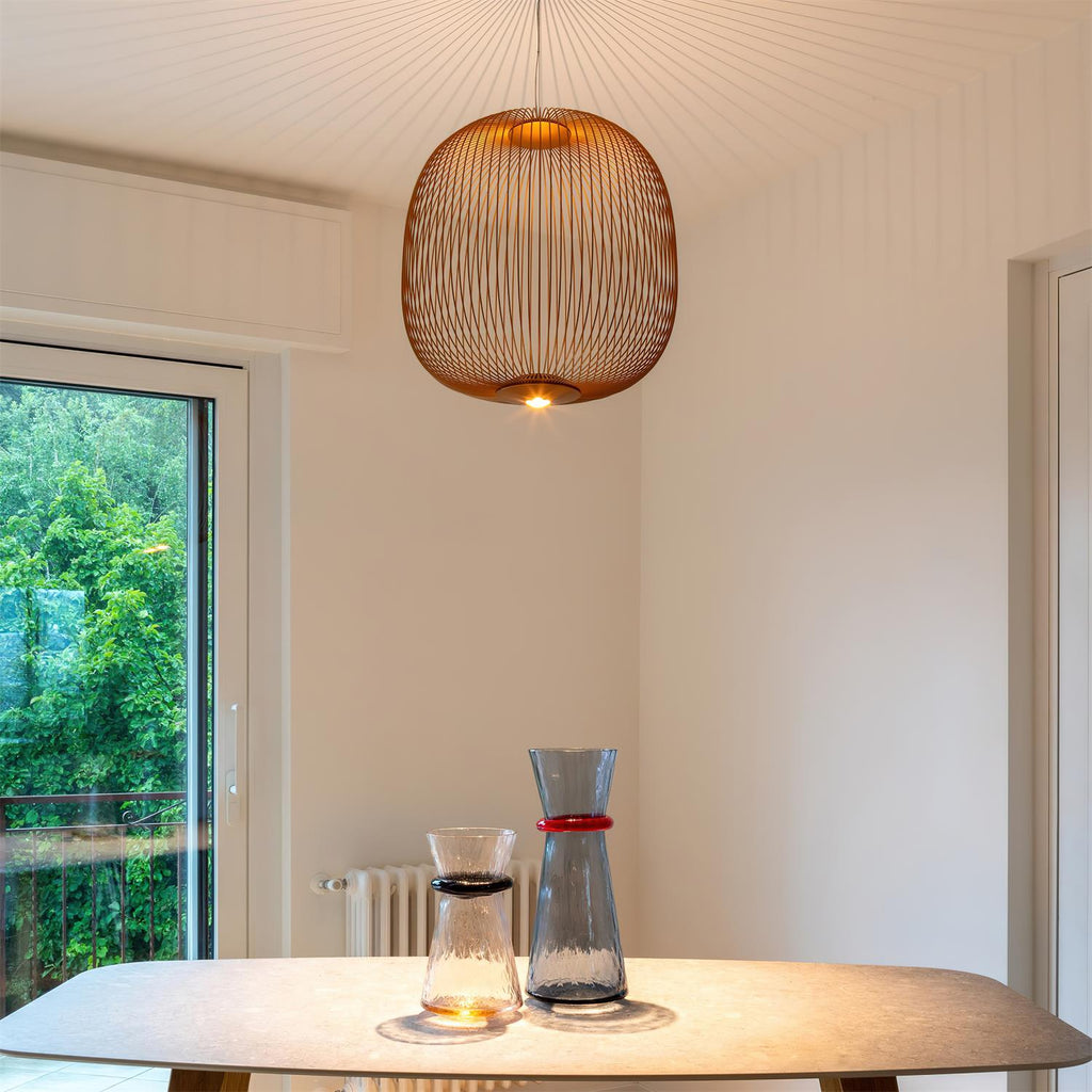 A copper-toned round Clérachic wire-cage pendant glows in a pale room beside a glass door, above a table with two glass carafes.