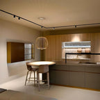 A pale wire-cage Clérachic pendant hangs over a round table beside a wood-fronted kitchen, under a track light on a warm cream ceiling.