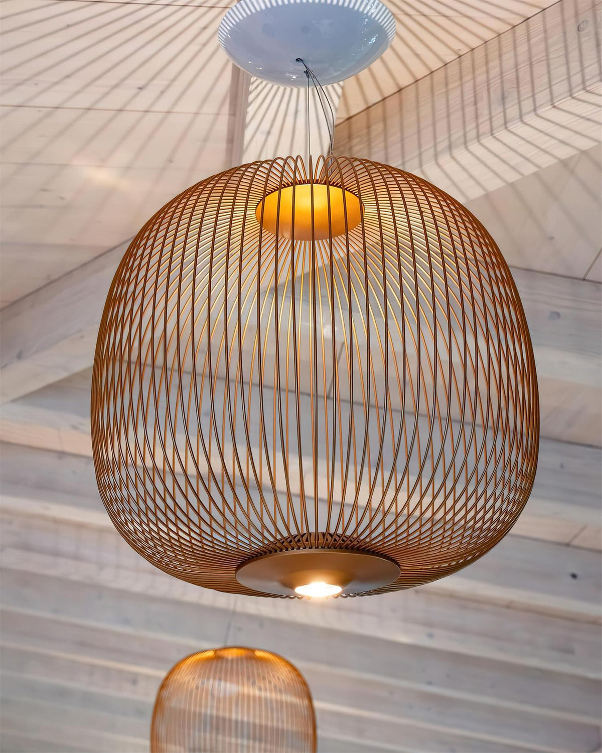Close view from below of a copper-toned Clérachic wire-cage pendant, its fine vertical wires and a lit base on a timber ceiling.