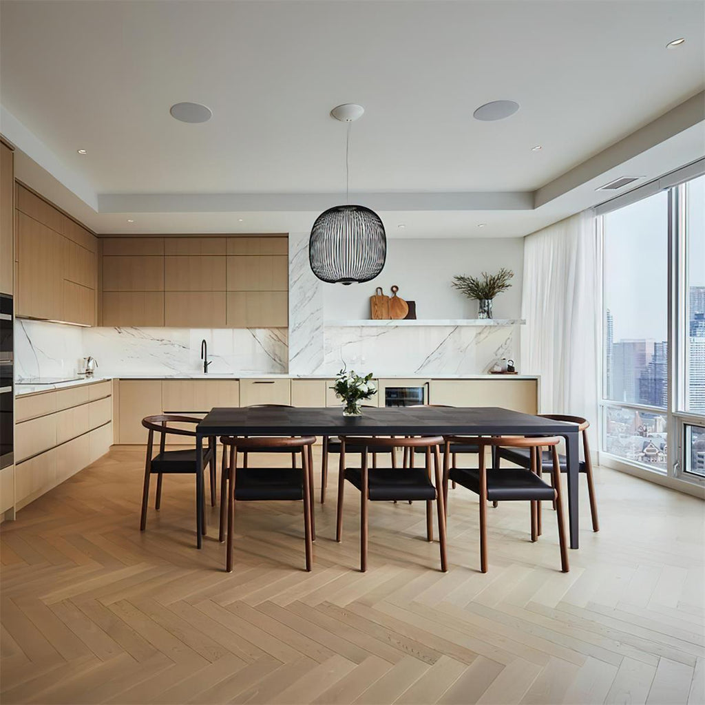A small black wire-cage Clérachic pendant hangs over a long dark dining table in a bright kitchen with a herringbone floor.