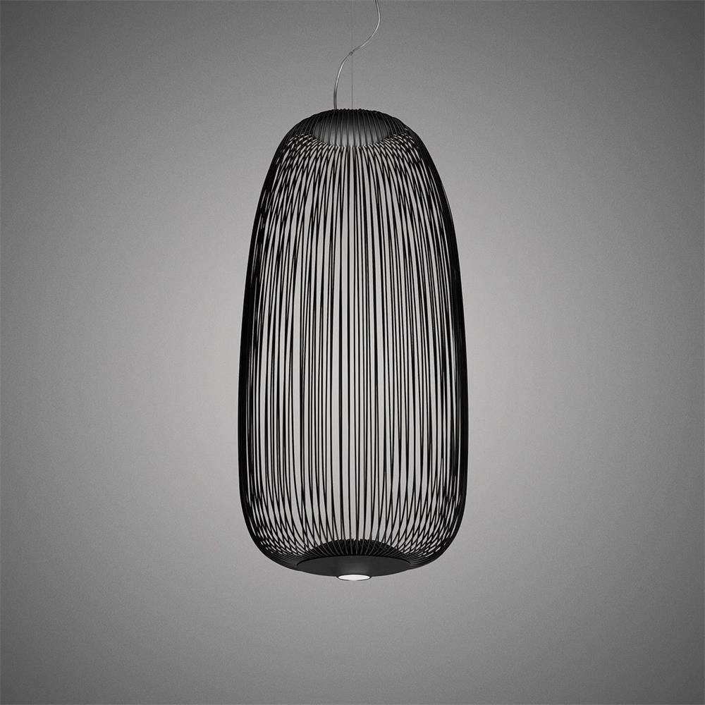 The tall oval wire-cage Clérachic pendant in black, with its fine wires and a lit base against a grey backdrop.