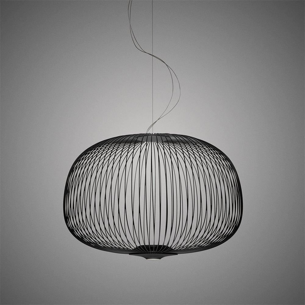 The low, wide drum-shaped wire-cage Clérachic pendant in black, with its curling cord above, on a dark grey backdrop.
